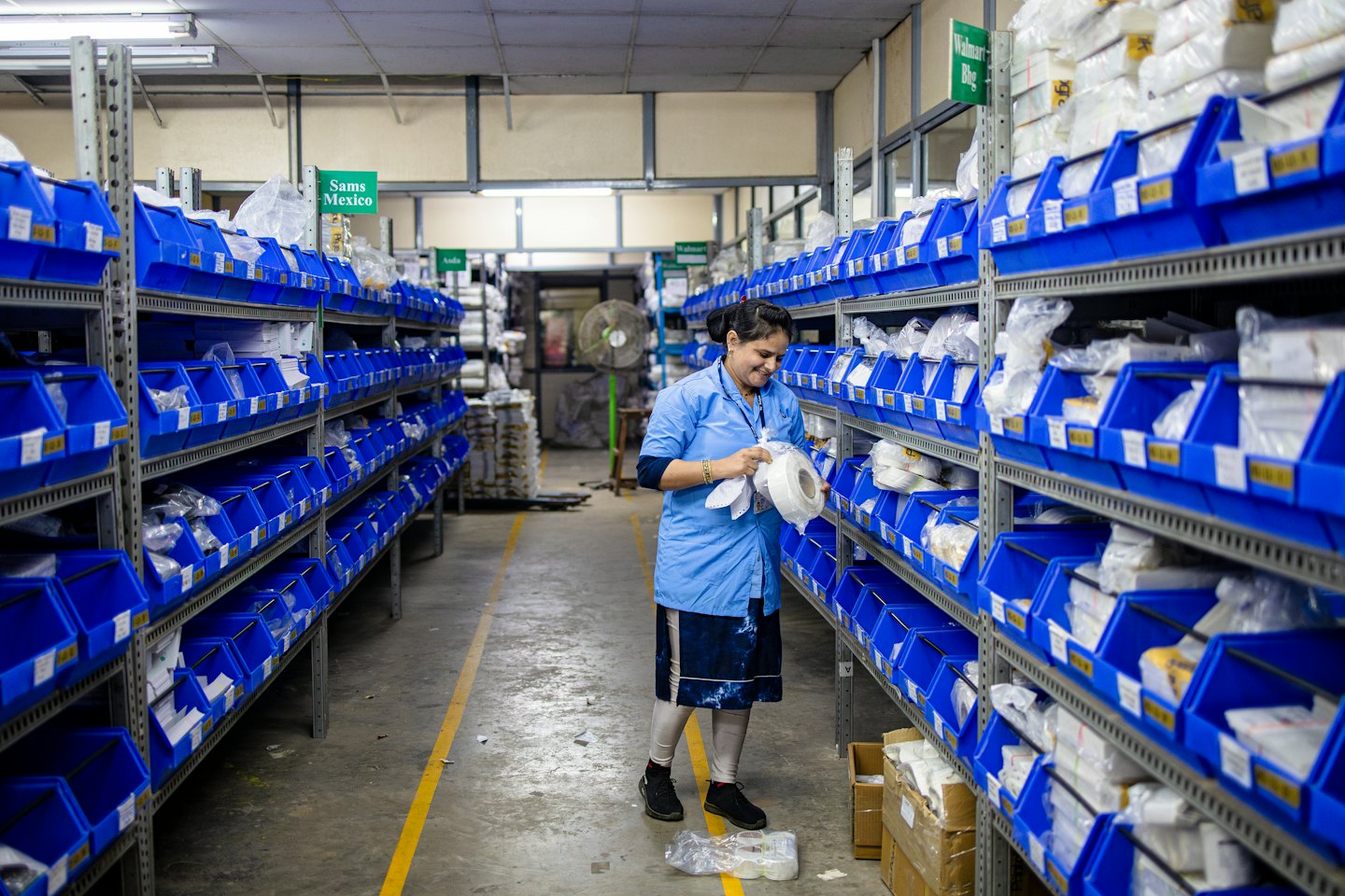 Woman in blue uniform sorts items from plastic bins in a warehouse.